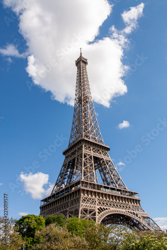 Obraz na plátně Eiffel Tower standing tall against a bright blue summer sky with a dramatic large white fluffy cloud centered behind its spire and green treetops below in Paris, Île-de-France, France