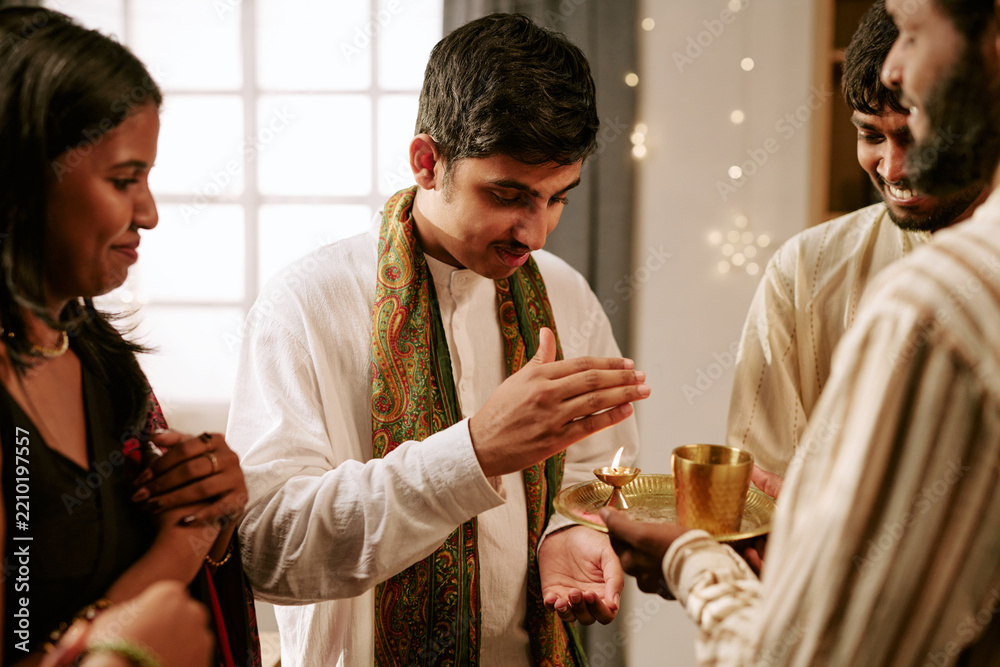 Obraz premium Indian young adult man warming hands over oil lamp flame during Diwali celebration with friends. Scene fits cultural marketing, religious holiday advertising, festive greeting campaigns