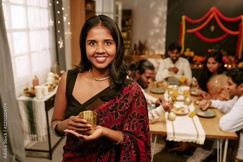 Obraz premium Portrait of smiling Indian young adult woman holding cup during festive dinner with family at home. Image suitable for holiday marketing campaigns and cultural celebration promotions