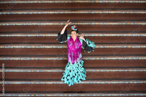 An Andalusian woman wearing a turquoise flamenco dress with black polka dots and a purple shawl, with flowers in her hair, dancing flamenco on a staircase in Seville’s Spain Square. Elevated view.