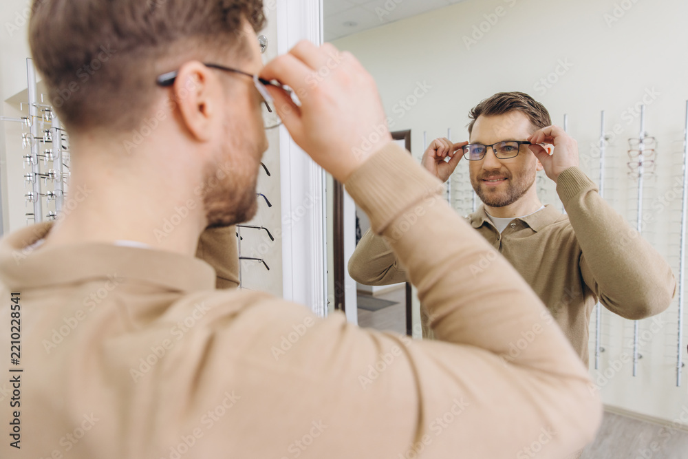 Obraz premium Man trying new frames in an optical shop, checking his look in the mirror for improved vision and style