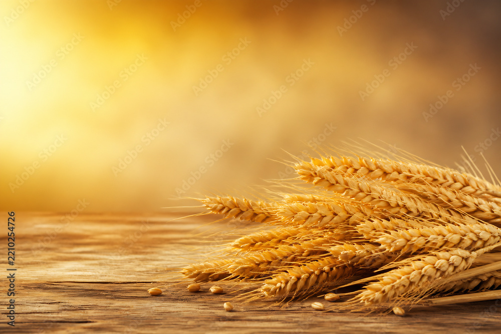 Naklejka premium A bunch of golden wheat stalks rests on a rustic wooden surface against a warm, blurred yellow and brown background.