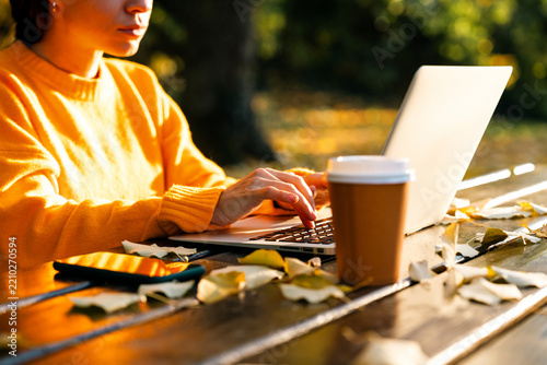 Modern woman working on laptop at picnic table in autumn park.