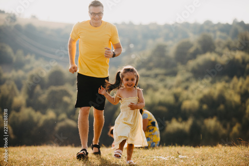 Father Chasing Daughter Through Sunlit Meadow During Summer Family Outdoor Playtime And Joyful Moments