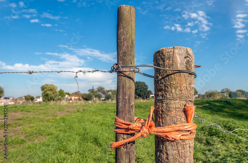 Close-up of rough wooden posts connected by iron wire and orange rope. The posts stand in the foreground of a rural area.