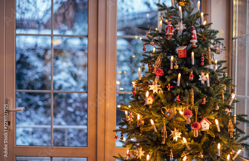 Christmas tree adorned with colorful ornaments and warm lights, standing near a window with a snowy landscape outside, creating a cozy holiday atmosphere