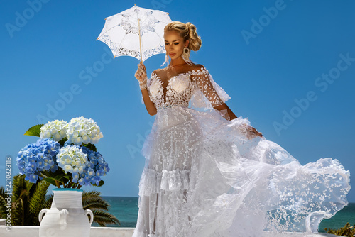Elegant Blonde Bride in Couture Lace Wedding Gown with Flying Veil and Parasol, Posing by Azure Sea with Blue Hydrangeas at a Luxury Mediterranean Coastal Resort