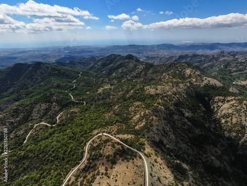 Aerial view of Kionia mountain peak with winding roads cutting through the lush green forests and rugged terrain under a bright blue sky in Lazanias, Lefkosia, Cyprus.