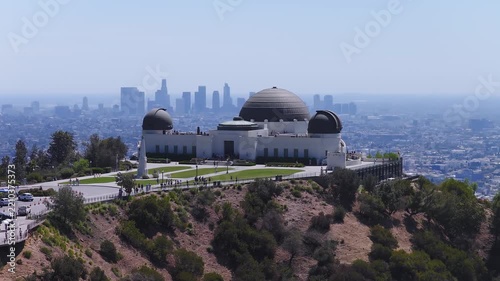 Aerial view shows Griffith Observatory situated on Mount Hollywood with visitors walking around the grounds and the downtown Los Angeles skyline in the distant haze.