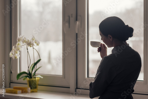 Silhouetted woman in dark uniform stands near window, holding small cup close while looking outside, soft natural backlight outlining profile, bun, and quiet interior mood with blurred winter view