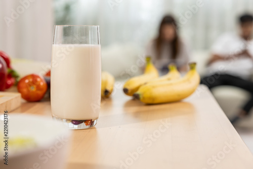 Milk glass on kitchen table with banana and fresh fruit, blurred family background creating warm breakfast scene