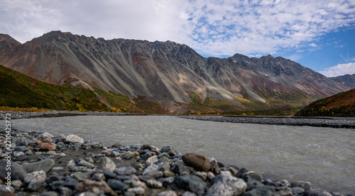 View of Rainbow Ridge with its rugged slopes, autumn foliage, and a glacial river flowing over rocky banks under a cloudy sky in Southeast Fairbanks Census Area, Alaska, United States.