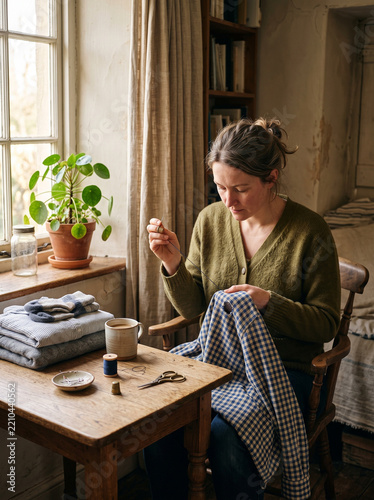 Woman mending a checkered shirt with needle and thread by a window. Female repairing clothes at home. Sustainable fashion and frugal lifestyle concept