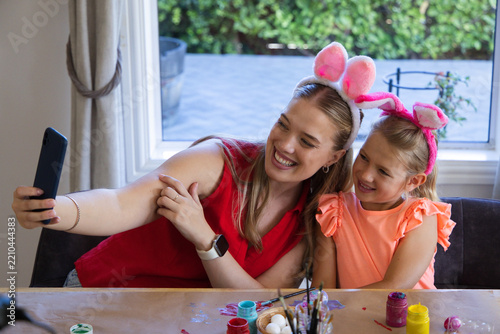 Mother and child posing with smartphone wearing bunny ears at table with paints and brushes