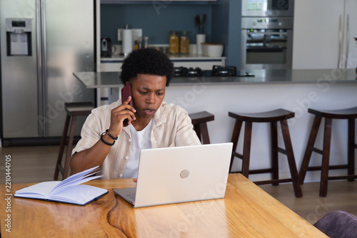 African man wearing light beige shirt holding smartphone and working on laptop at dining table