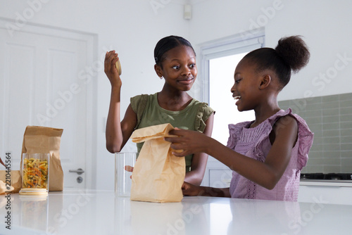 African American sisters, teen and child, standing at white counter handling paper bags and jar