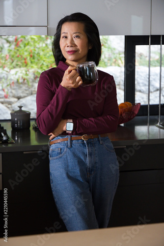Mature Asian woman standing in modern kitchen holding glass mug wearing burgundy top and smartwatch