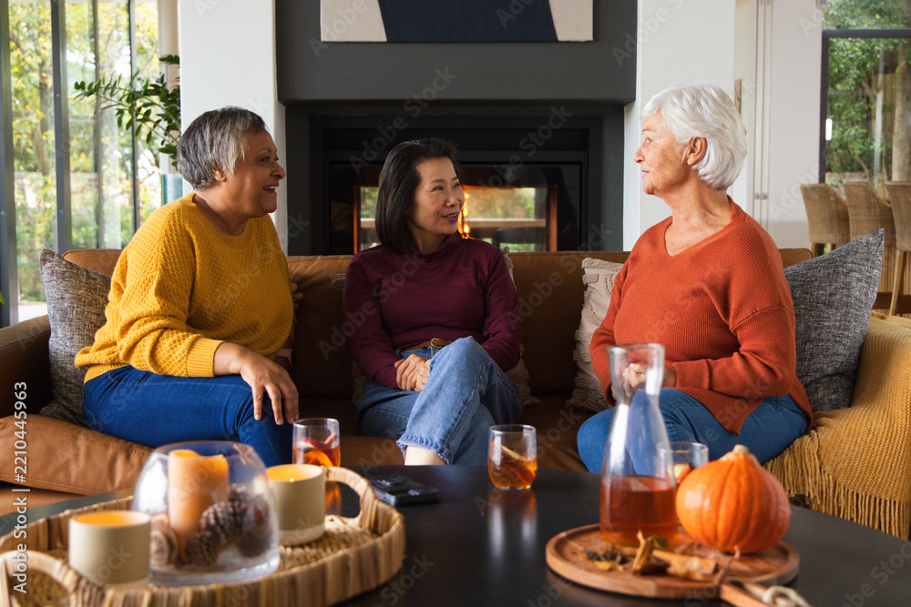 Fototapeta premium Diverse female friends in sweaters and jeans chatting and sipping amber drinks on sofa by fireplace