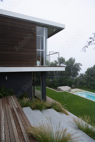 Modern two-story house sitting on sloped lot with wooden deck, balcony, concrete steps, pool