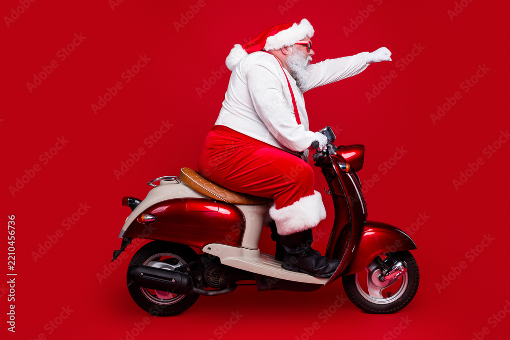 Obraz premium Santa Claus riding a red vintage scooter in festive costume on a red background, playful holiday transportation and Christmas celebration scene