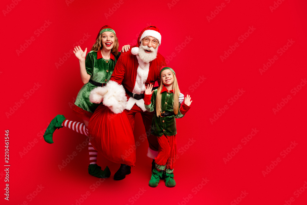 Obraz premium Santa Claus with two cheerful elves posing with a large red gift sack in festive green and red costumes — family holiday portrait conveying joyful Christmas celebration