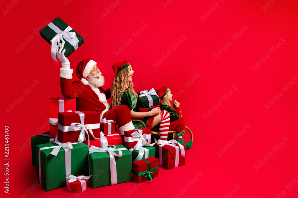 Obraz premium Santa Claus and two festive elves sitting among wrapped Christmas presents on a red background, celebrating holiday joy, family fun and playful seasonal cheer