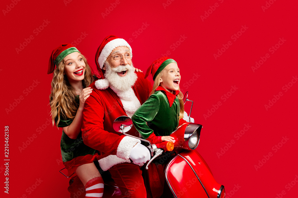 Obraz premium Santa Claus with two cheerful elves riding a red scooter, laughing and celebrating Christmas together in a joyful family holiday scene full of festive fun and seasonal tradition