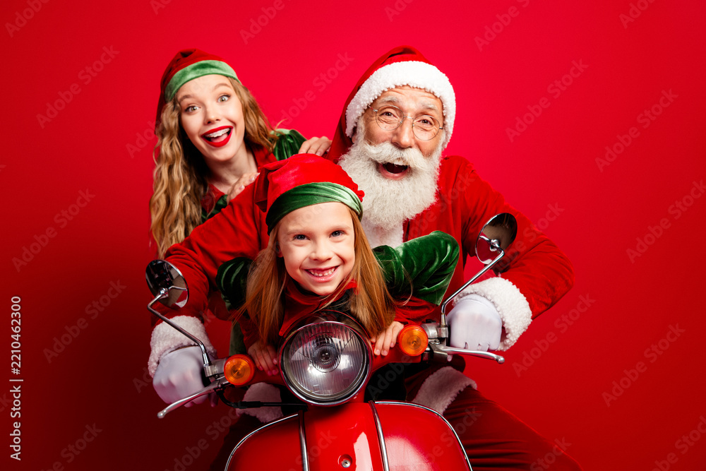 Obraz premium Santa and two cheerful elves riding a red scooter for Christmas, smiling family holiday portrait with festive hats and joyful expressions