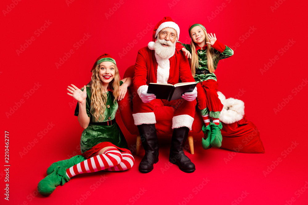 Obraz premium Santa Claus reading with two cheerful elves and children on a red background for Christmas holiday family portrait and festive storytelling