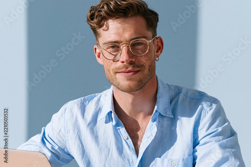 Young man with glasses sitting beside laptop in soft daylight, relaxed and focused expression, wearing light blue shirt and earring