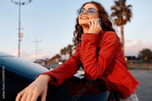 Young woman in a red jacket smiling while talking on her phone outdoors. She leans against a car during sunset. Lifestyle concept for modern communication, travel, and urban lifestyle.