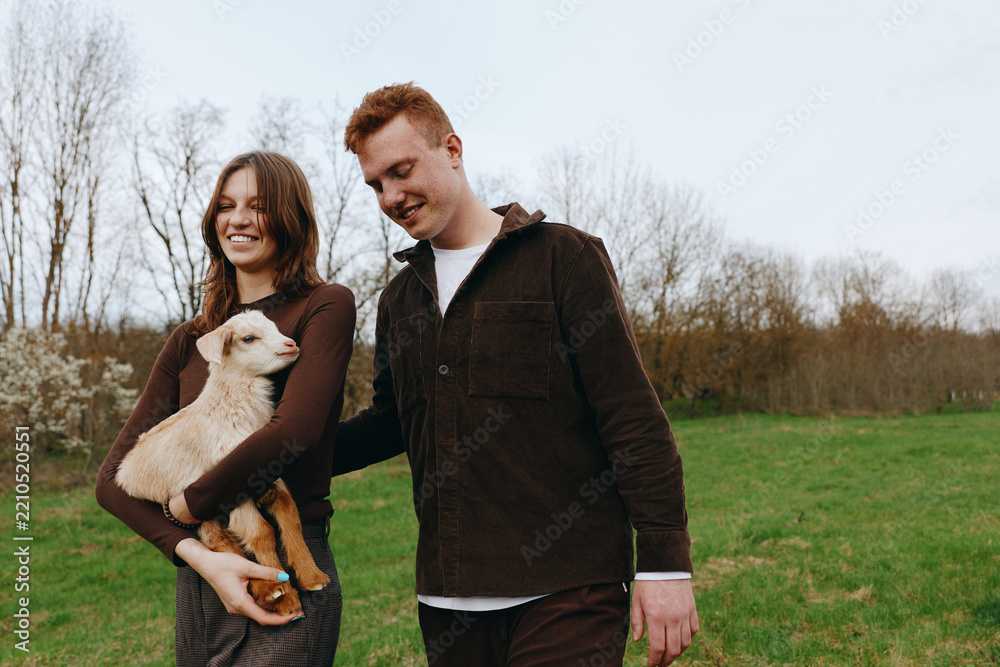 Obraz premium Joyful couple outdoors with a goat kid, walking through a green field, warm light, smiling faces, relaxed posture, natural tones, carefree moment, countryside vibe, gentle breeze vibrant