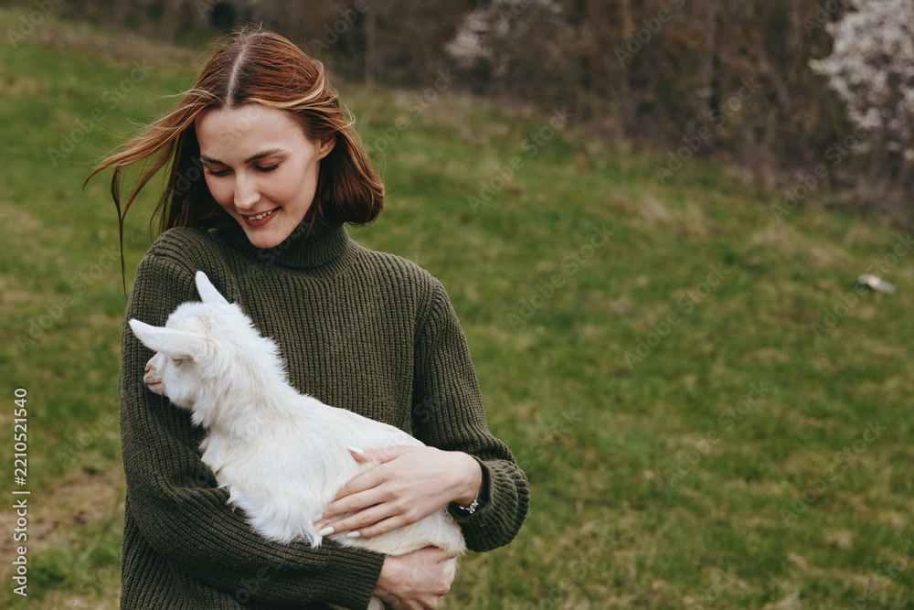 Obraz premium Woman in a chunky knitted sweater hugs a white goat kid in a grassy field. Natural light, candid lifestyle moment, warm mood, gentle affection between human and animal in an outdoor nature scene.