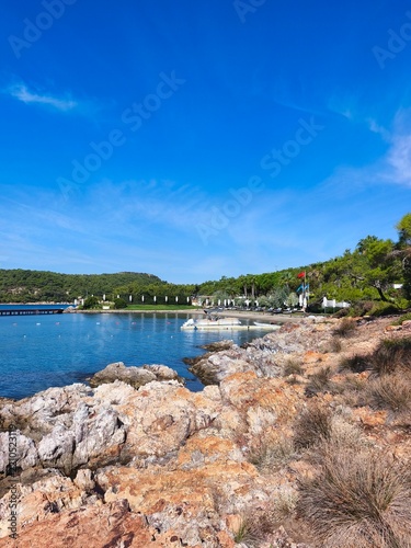 Scenic view of Ortunc Cove in Cunda Island, Ayvalik with crystal clear sea, pebble beach, pine trees, and a small boat under a blue sky, Balikesir, Turkey.