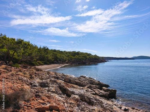 Scenic view of Ortunc Cove in Cunda Island, Ayvalik with crystal clear sea, pebble beach, pine trees, and a small boat under a blue sky, Balikesir, Turkey.