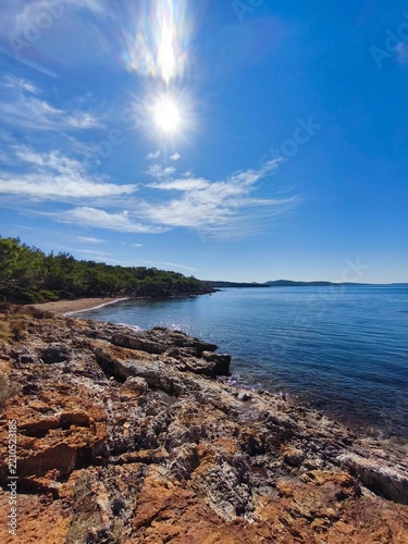 Scenic view of Ortunc Cove in Cunda Island, Ayvalik with crystal clear sea, pebble beach, pine trees, and a small boat under a blue sky, Balikesir, Turkey.