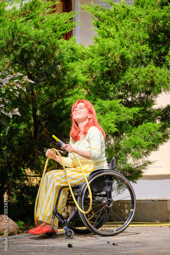 Smiling woman with disability sitting in wheelchair and holding garden hose outdoors. Happy female wheelchair user gardening on a sunny summer day.
