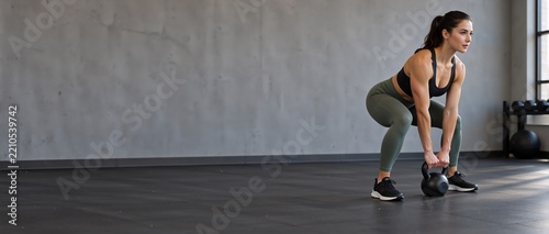 Young athletic Caucasian woman performing kettlebell deadlift exercise in modern gym, strength training, fitness workout, functional movement, focused, athletic apparel, concrete wall and gym