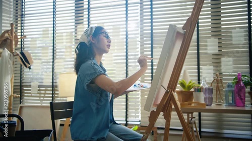 Gen Z Asian woman freelance artist painting with paintbrush on canvas easel, representing creative professional women designer, online independent creator and art studio hobby lifestyle