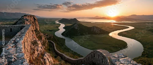 Panel kuchenny z motywem Bucketlist wanderlust from atop a medieval fortress wall a serpentine river curves through emerald floodplains toward Lake Skadar at golden sunset, mountains glowing on the horizon.
