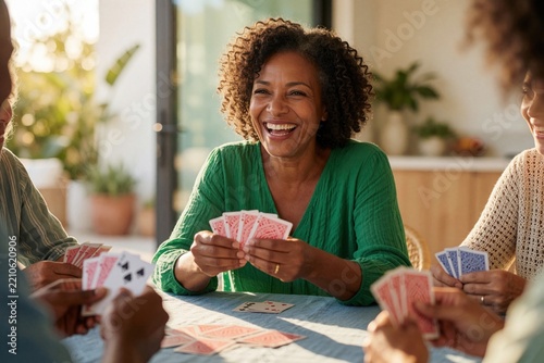 Smiling middle aged Black woman plays cards with diverse friends at home. Happy African American lady holds playing cards during fun game night. Group of people laugh, enjoy leisure time together at