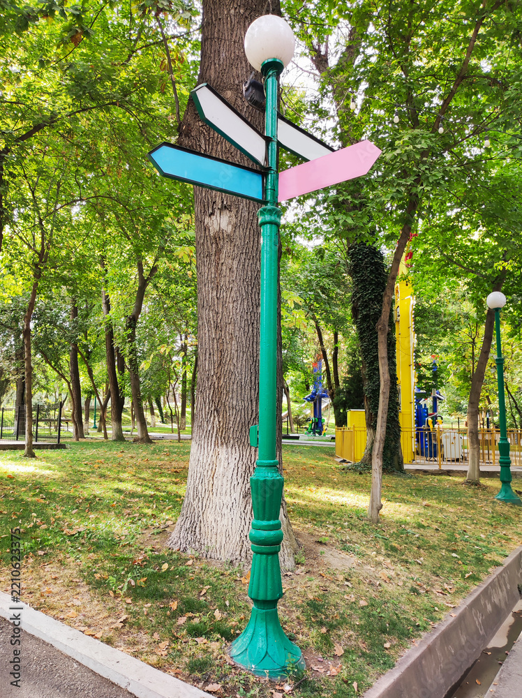 Fototapeta premium Green vintage lamppost with blank colorful directional arrow signs standing near large tree trunk in sunny city park. Navigation, wayfinding and urban park infrastructure concept.