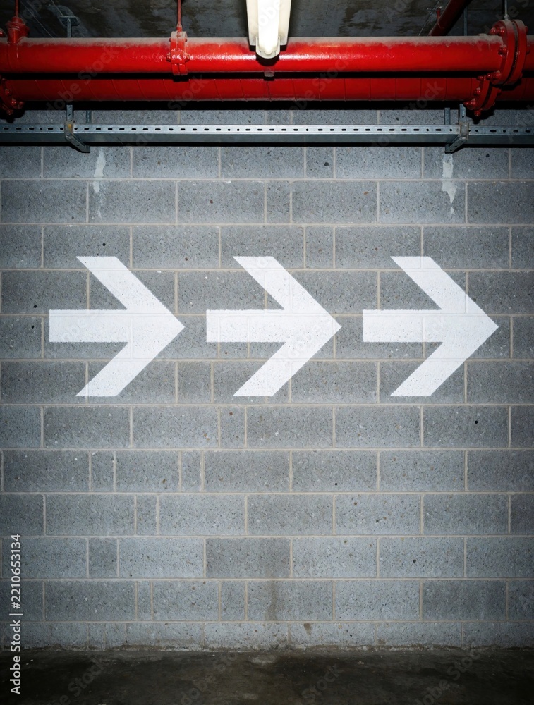 Fototapeta premium Three white arrow symbols painted on gray cinder block wall. Directional signs point right in industrial building underground parking lot. Concrete wall with red pipes above shows way to exit