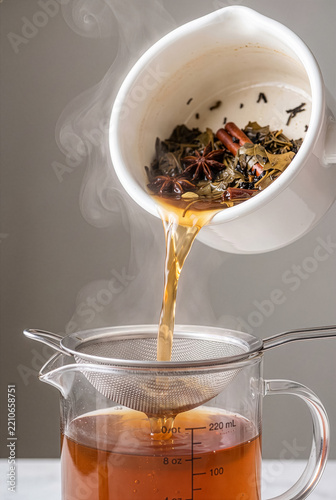 Pouring hot spiced chai tea through strainer into glass measuring jug