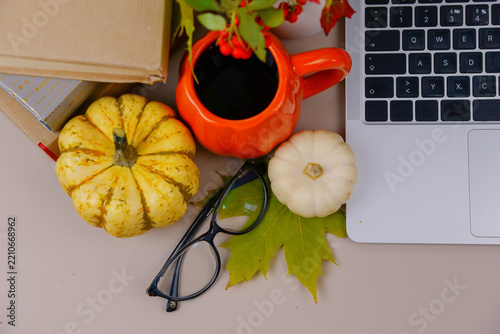Cozy autumn workspace with laptop, pumpkins, coffee, glasses, books and colorful fall leaves arranged on a neutral desk.