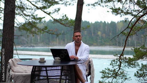 Successful businessman in bathrobe typing on laptop while working remotely on a foggy riverbank terrace in autumn.