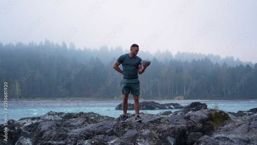 Fototapeta premium Front view of a strong young male tourist practicing strength workout and lifting a rock on a misty morning in Altai