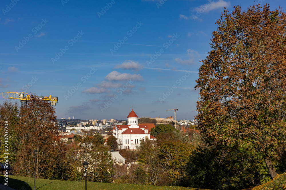 Fototapeta premium Panoramic autumn view of Vilnius, Lithuania, on a sunny October day, featuring a historic white church with red tiled roofs surrounded by colorful autumn trees, with construction cranes.