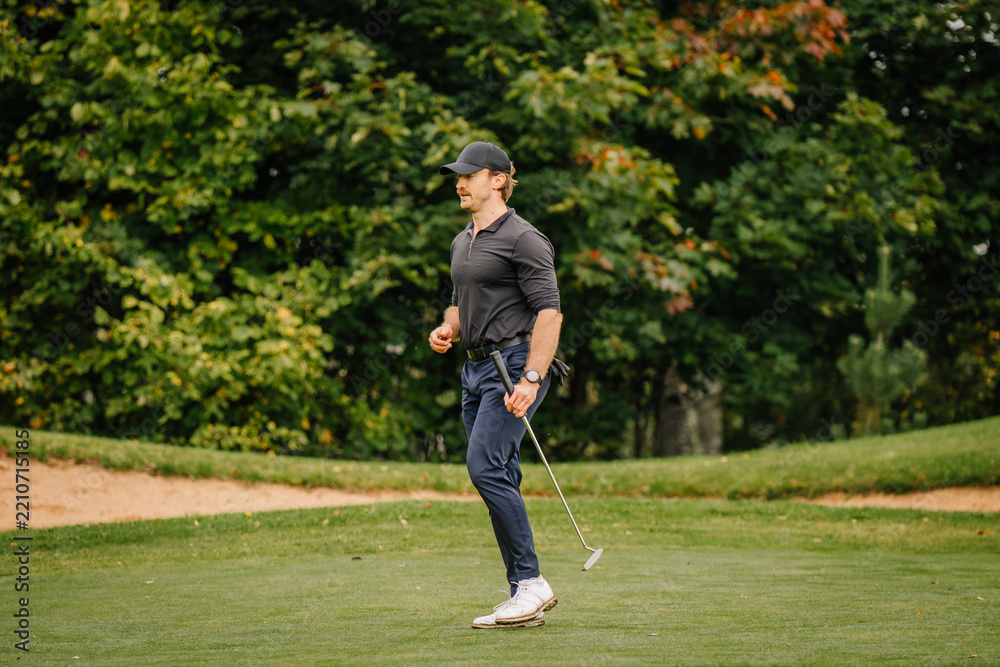 Obraz premium Golfer walking across manicured grass holding a club beside a sandy path and lush green trees in an outdoor recreational landscape scene