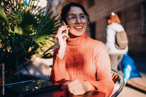 Happy hispanic woman phoning with smartphone and smiling, joyful mobile conversation in city cafe, looking away, authentic representation, positive attitude, connection and digital lifestyle.